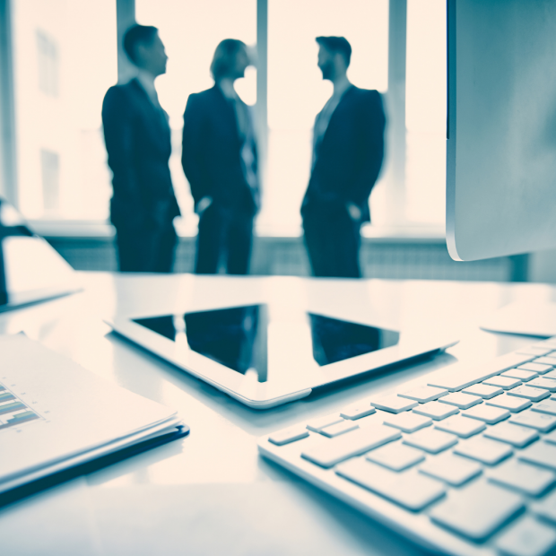 three men in the background by a window, computer and ipad in foreground, black and white picture